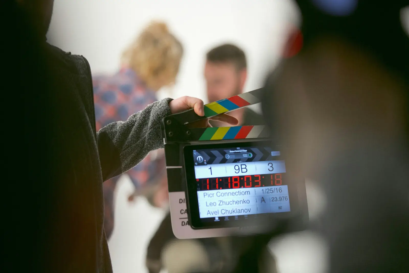 Filmklappe vor einer Szene am Set, im Hintergrund unscharf zwei Darsteller