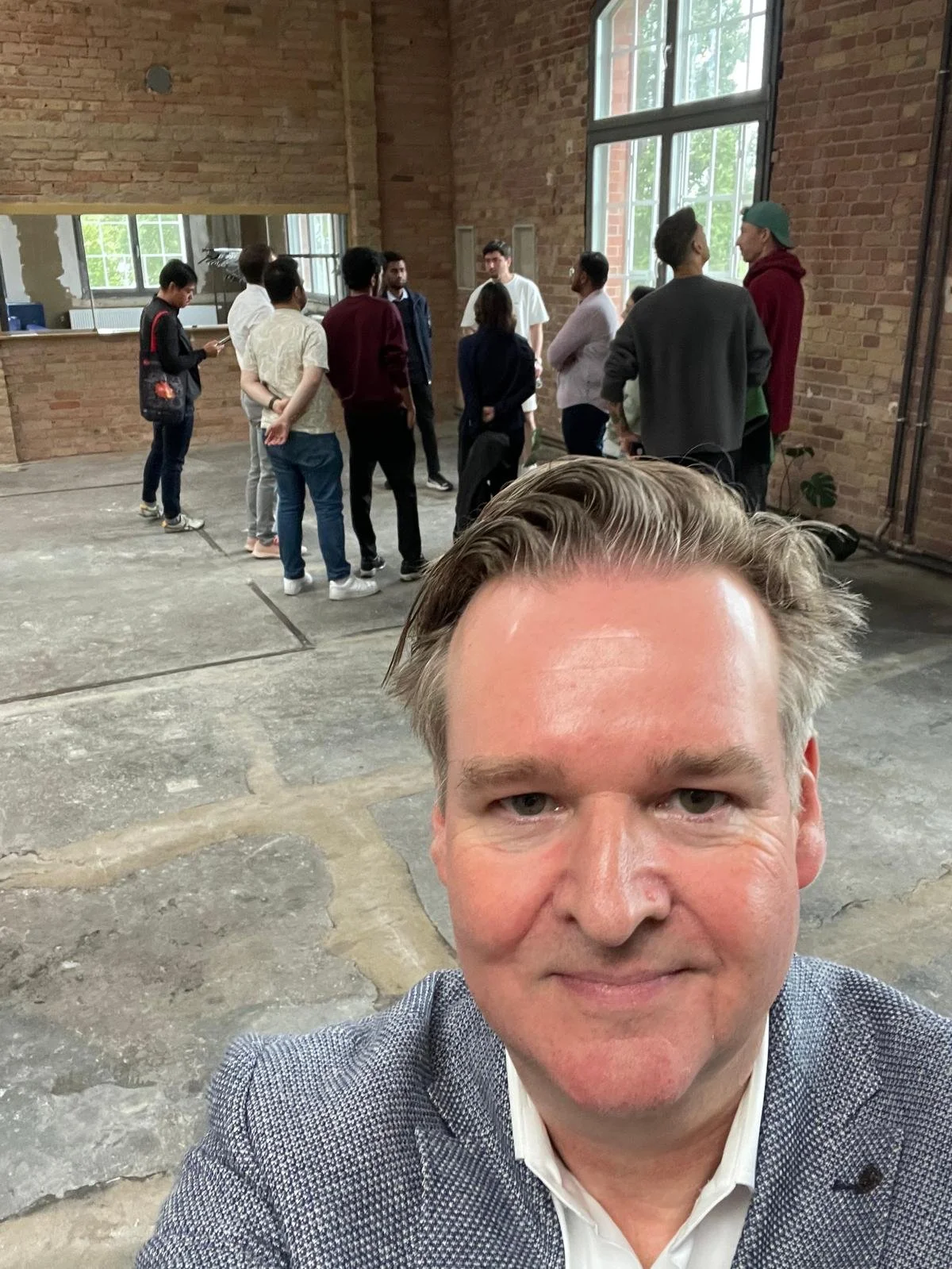Christian Simanek in einer leeren Industriehalle mit Backsteinwänden, dahinter eine Gruppe Studierender